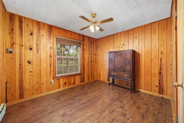 empty room featuring wooden walls, ceiling fan, dark hardwood / wood-style floors, and a baseboard heating unit