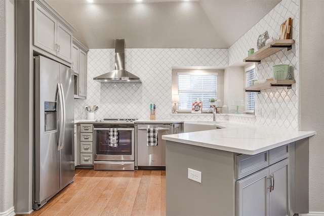 kitchen with kitchen peninsula, wall chimney range hood, gray cabinets, stainless steel appliances, and light hardwood / wood-style floors