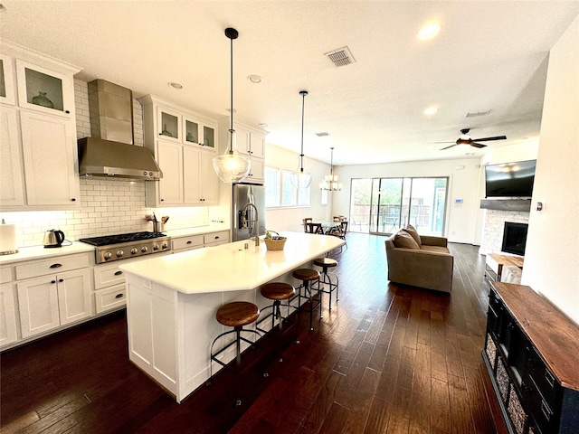 kitchen with wall chimney range hood, white cabinets, an island with sink, dark hardwood / wood-style floors, and appliances with stainless steel finishes