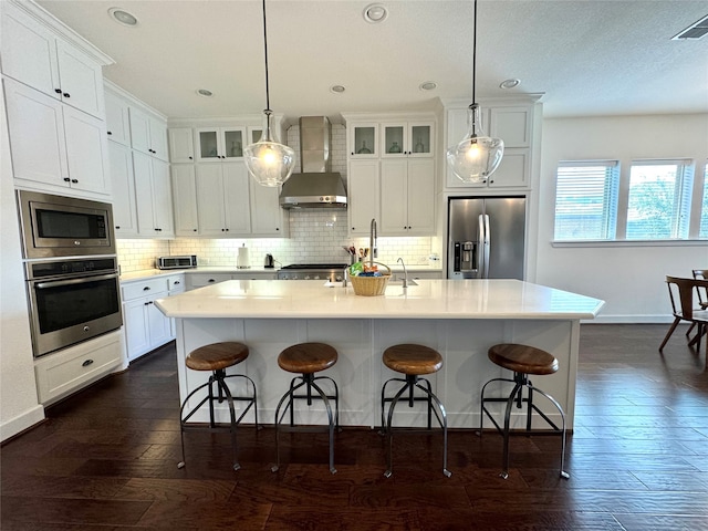 kitchen with an island with sink, wall chimney exhaust hood, dark hardwood / wood-style floors, appliances with stainless steel finishes, and backsplash