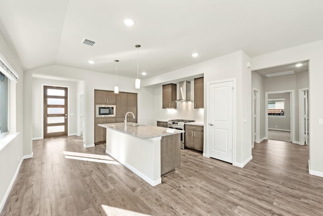 kitchen featuring stainless steel appliances, wall chimney range hood, decorative light fixtures, a center island with sink, and light wood-type flooring