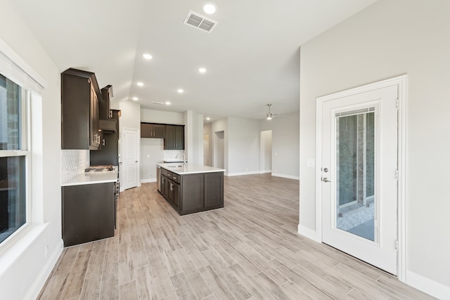 kitchen with dark brown cabinets, backsplash, ceiling fan, a kitchen island with sink, and light hardwood / wood-style flooring