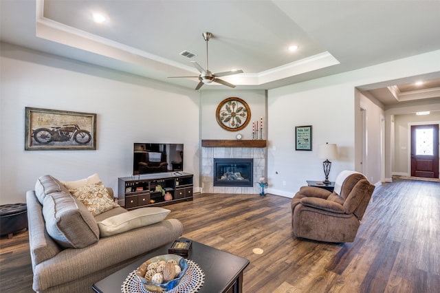 living room with dark hardwood / wood-style floors, a tiled fireplace, ornamental molding, ceiling fan, and a raised ceiling