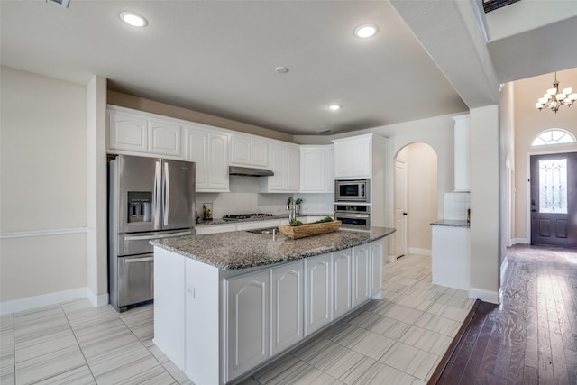 kitchen with stainless steel appliances, a kitchen island with sink, an inviting chandelier, dark stone countertops, and white cabinets