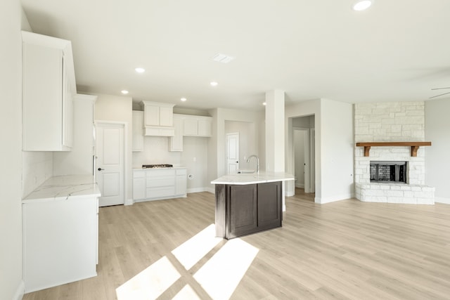 kitchen featuring backsplash, a stone fireplace, light hardwood / wood-style floors, white cabinets, and a kitchen island with sink