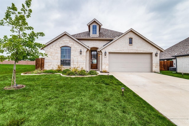 french country home with roof with shingles, central air condition unit, concrete driveway, an attached garage, and a front lawn