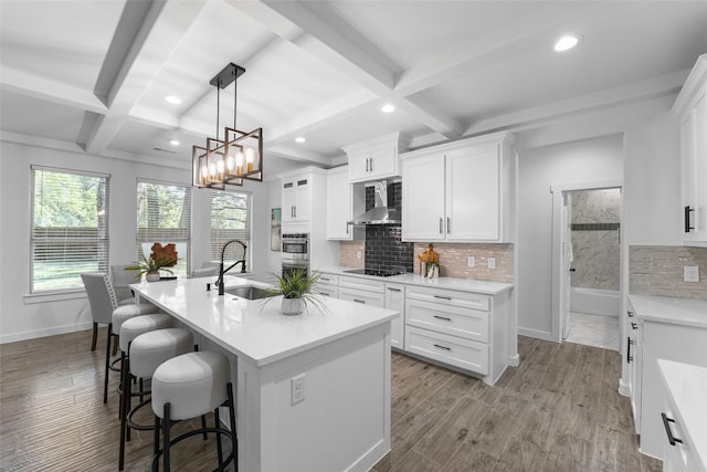 kitchen featuring wall chimney range hood, sink, a center island with sink, beamed ceiling, and white cabinetry