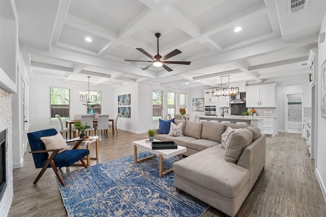 living room featuring coffered ceiling, ceiling fan with notable chandelier, sink, light hardwood / wood-style floors, and a stone fireplace