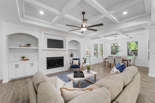 living room featuring built in shelves, light hardwood / wood-style floors, a stone fireplace, and ceiling fan with notable chandelier