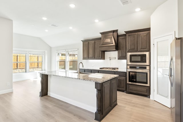 kitchen with sink, custom exhaust hood, a kitchen island with sink, light stone counters, and stainless steel appliances