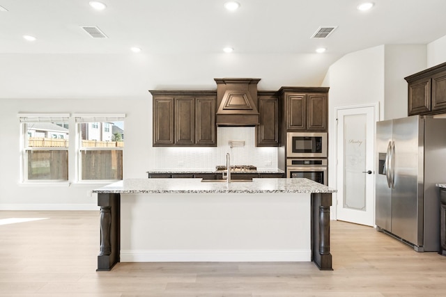 kitchen featuring dark brown cabinetry, light stone counters, stainless steel appliances, a kitchen island with sink, and decorative backsplash