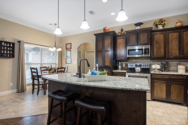 kitchen with appliances with stainless steel finishes, light stone counters, dark brown cabinetry, decorative light fixtures, and an island with sink