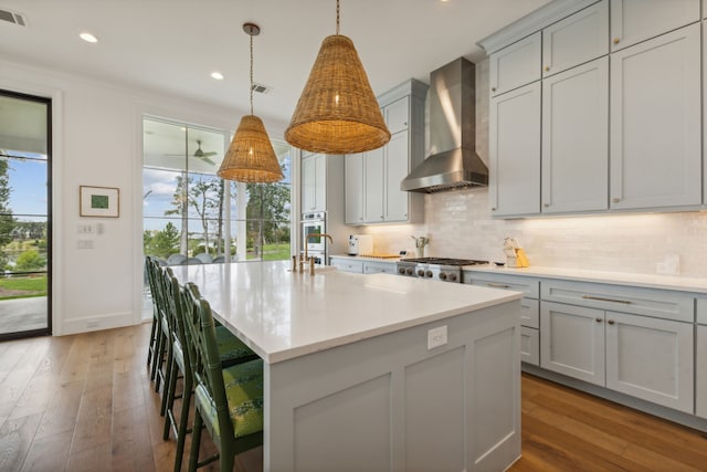 kitchen with a center island with sink, wall chimney exhaust hood, backsplash, wood-type flooring, and pendant lighting