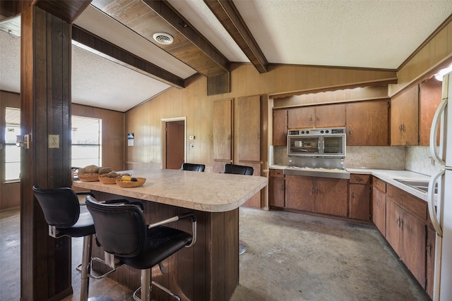 kitchen with stainless steel oven, vaulted ceiling with beams, a textured ceiling, wooden walls, and a kitchen bar