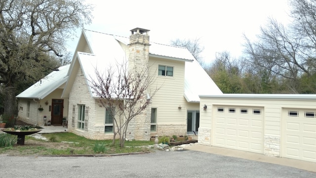 view of side of home featuring a garage, stone siding, a chimney, metal roof, and driveway
