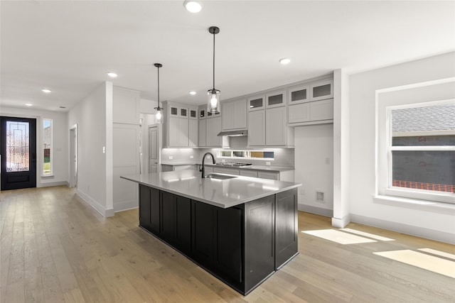 kitchen featuring decorative backsplash, hanging light fixtures, sink, an island with sink, and light hardwood / wood-style flooring