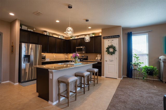 kitchen with decorative light fixtures, stainless steel appliances, light stone counters, a center island with sink, and light tile patterned floors