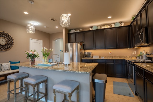 kitchen with light tile patterned floors, light stone counters, a kitchen island with sink, hanging light fixtures, and appliances with stainless steel finishes