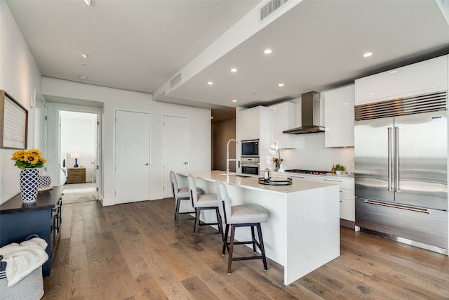 kitchen featuring white cabinetry, wall chimney exhaust hood, built in appliances, and a center island with sink