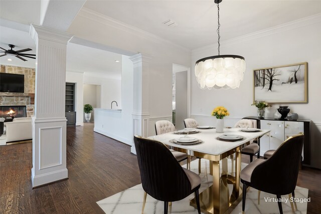 dining space featuring decorative columns, ceiling fan with notable chandelier, dark hardwood / wood-style flooring, a stone fireplace, and ornamental molding