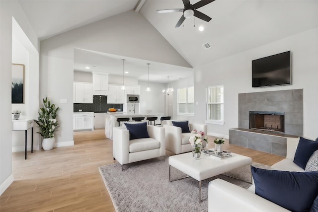 living room with a tiled fireplace, ceiling fan with notable chandelier, high vaulted ceiling, and light wood-type flooring