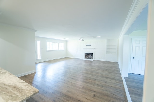 unfurnished living room featuring dark wood-type flooring, crown molding, a brick fireplace, built in shelves, and ceiling fan