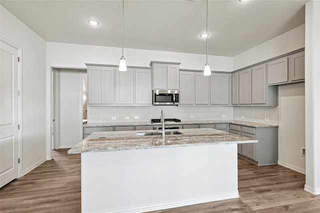 kitchen with light stone counters, dark hardwood / wood-style flooring, a kitchen island with sink, pendant lighting, and stainless steel appliances