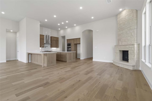 kitchen with light hardwood / wood-style flooring, wall chimney range hood, stainless steel appliances, a stone fireplace, and a kitchen bar