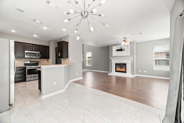 kitchen with decorative backsplash, light stone countertops, dark brown cabinets, ceiling fan with notable chandelier, and stainless steel appliances