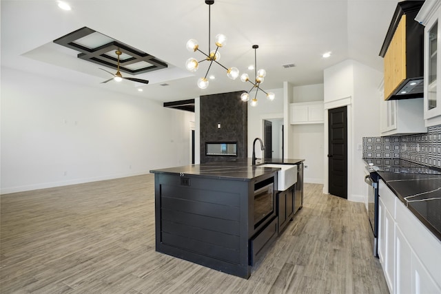 kitchen with sink, light hardwood / wood-style floors, ceiling fan with notable chandelier, an island with sink, and coffered ceiling