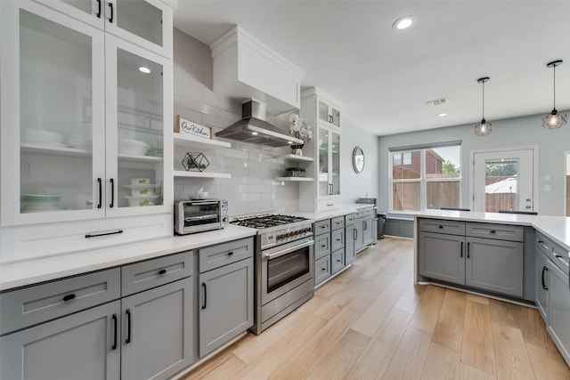 kitchen with wall chimney range hood, backsplash, stainless steel stove, gray cabinets, and hanging light fixtures