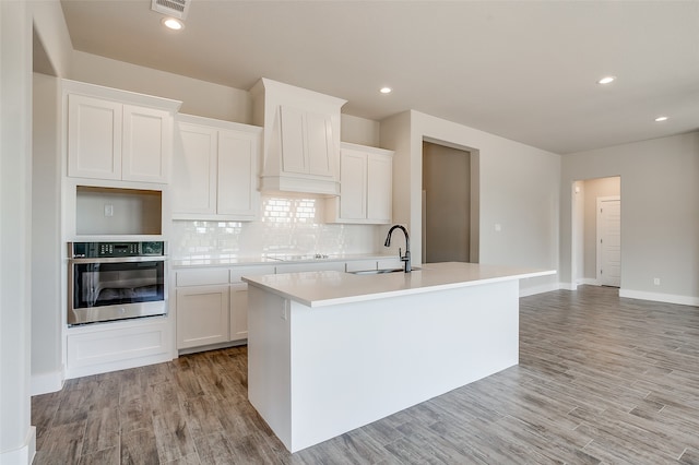 kitchen featuring light hardwood / wood-style floors, white cabinetry, stainless steel oven, a kitchen island with sink, and sink