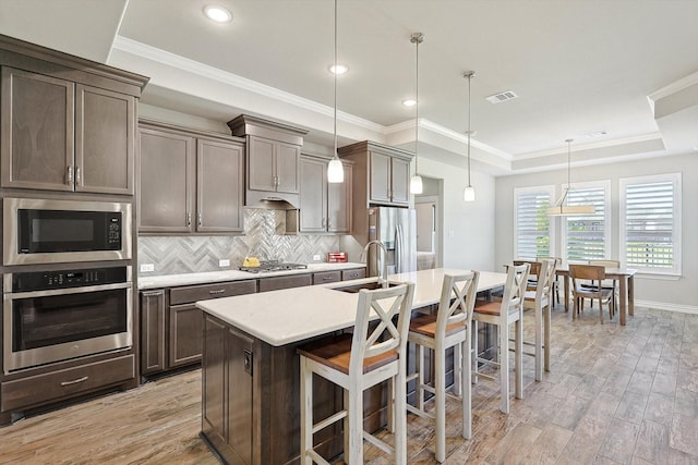 kitchen with appliances with stainless steel finishes, an island with sink, a kitchen bar, decorative light fixtures, and a raised ceiling