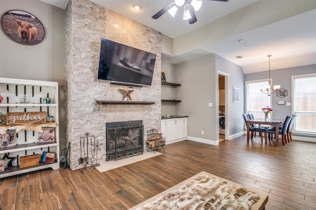 living room featuring washer / clothes dryer, dark wood-type flooring, ceiling fan with notable chandelier, and a stone fireplace