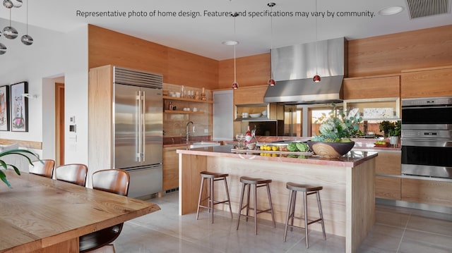 kitchen with pendant lighting, wall chimney exhaust hood, a kitchen bar, appliances with stainless steel finishes, and light tile patterned floors