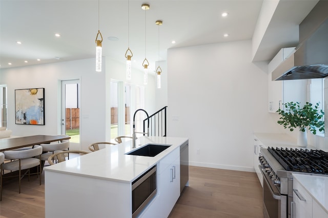 kitchen featuring stainless steel appliances, wall chimney range hood, sink, an island with sink, and wood-type flooring