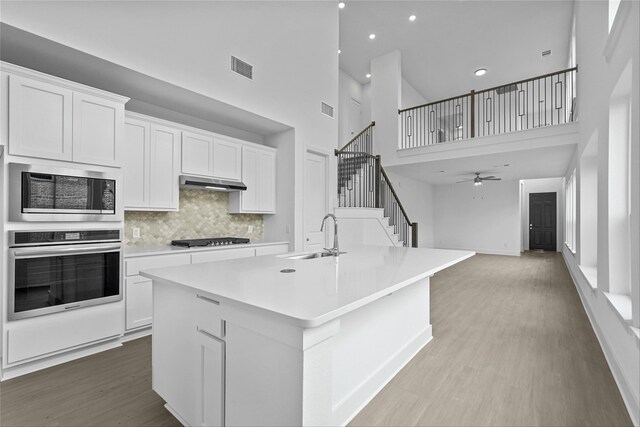 kitchen with a high ceiling, a center island with sink, sink, appliances with stainless steel finishes, and white cabinetry