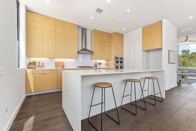 kitchen with dark hardwood / wood-style floors, backsplash, light brown cabinets, and wall chimney range hood