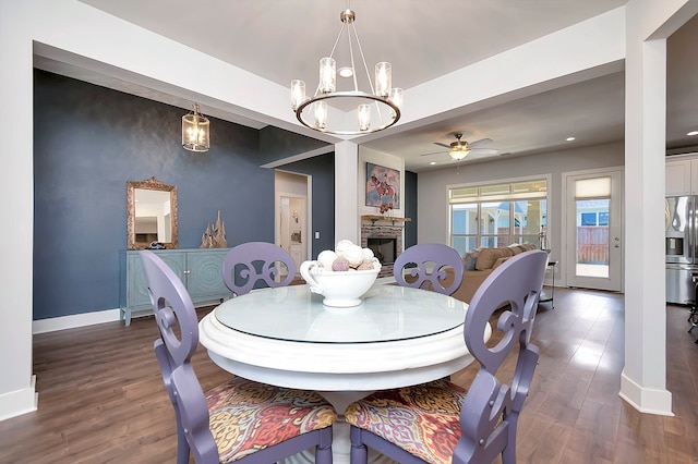 dining room with a stone fireplace, ceiling fan with notable chandelier, and dark hardwood / wood-style flooring