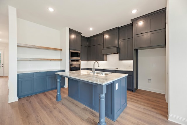 kitchen featuring light stone countertops, appliances with stainless steel finishes, an island with sink, sink, and light wood-type flooring