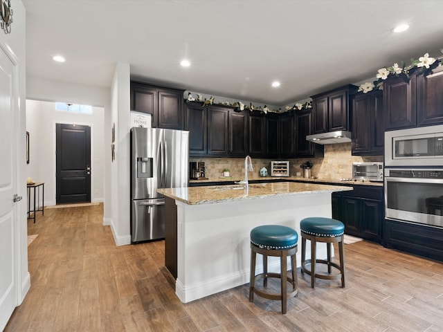 kitchen featuring decorative backsplash, a center island with sink, light wood-type flooring, stainless steel appliances, and light stone counters