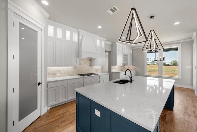 kitchen featuring light wood-type flooring, premium range hood, backsplash, an island with sink, and crown molding