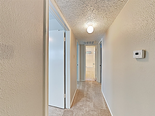 corridor with a textured ceiling, a textured wall, light carpet, visible vents, and baseboards