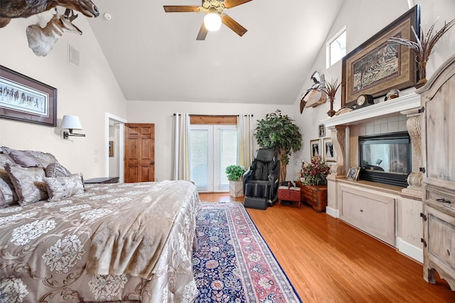 bedroom with ceiling fan, a tiled fireplace, light hardwood / wood-style flooring, and high vaulted ceiling