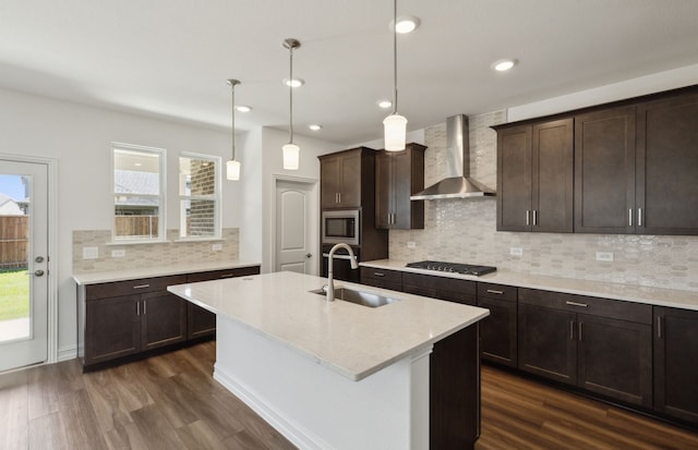 kitchen with wall chimney range hood, sink, stainless steel appliances, light stone counters, and a center island with sink