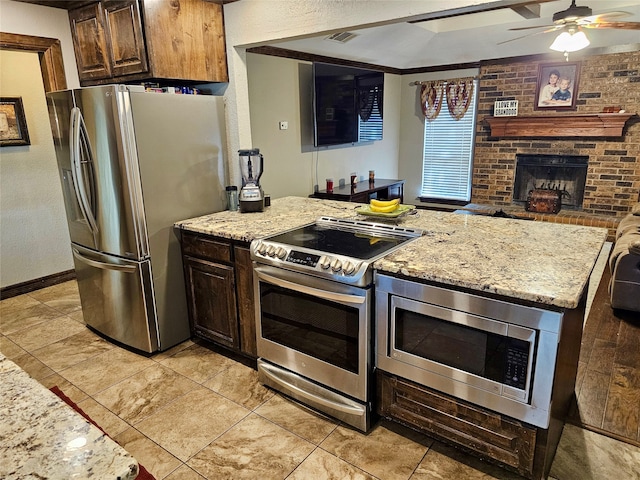 kitchen featuring stainless steel appliances, ceiling fan, a brick fireplace, and light stone counters