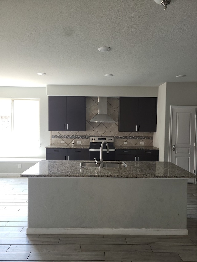 kitchen with stone counters, tasteful backsplash, a center island with sink, and wall chimney range hood