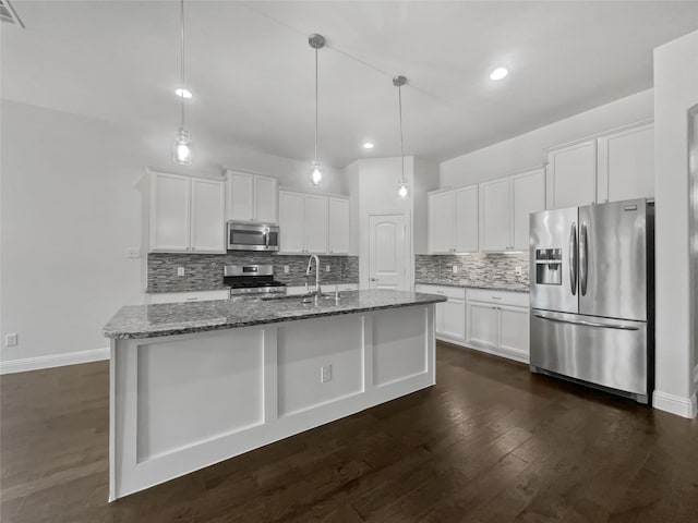 kitchen featuring dark stone counters, stainless steel appliances, a center island with sink, white cabinets, and hanging light fixtures