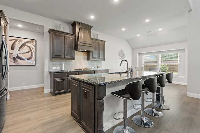 kitchen with custom exhaust hood, sink, a center island with sink, light hardwood / wood-style flooring, and backsplash