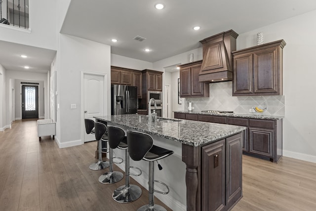 kitchen featuring premium range hood, stainless steel appliances, decorative backsplash, a center island with sink, and light hardwood / wood-style floors